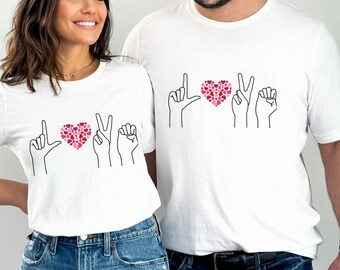May include: Two white t-shirts with a graphic of hands forming the word "LOVE" in American Sign Language. The heart in the graphic is made up of small pink and red hearts.