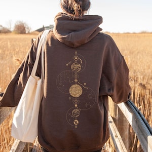 Puede incluir: Una sudadera con capucha marrón con un diseño celestial en la espalda. El diseño presenta planetas, estrellas y otros elementos celestiales en color dorado. La sudadera tiene una capucha con cordón y un bolsillo delantero.
