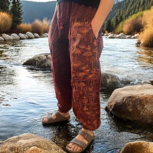 May include: Deep red loose-fitting trousers featuring a repeating pattern of mushrooms and butterflies in a lighter orange hue. The pants include a drawstring waist, side pockets, and a horizontal zippered pocket on the thigh with orange trim detailing.