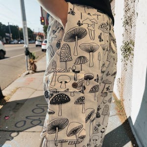 May include: Off-white cargo pants featuring a repeating black line-art pattern of various mushrooms and small butterflies. The pants include a large side cargo pocket with a single brown button closure on the flap and a side hand pocket.