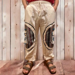 May include: Cream-colored pants with decorative patches featuring a mix of patterns and colors, including blue, red, and black. The pants have a drawstring waist and pockets. The person is wearing brown sandals.