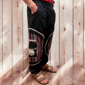 May include: Black pants with decorative patterned panels and pockets. The panels feature a mix of red, white, and brown geometric designs. The pants have a drawstring waist and are worn with brown sandals.