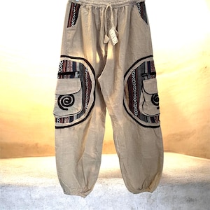 May include: Tan-colored, loose-fitting pants with a drawstring waist and two cargo pockets. The pockets and side panels feature a colorful, patterned design with red, blue, and black accents. The pants have a relaxed, bohemian style.