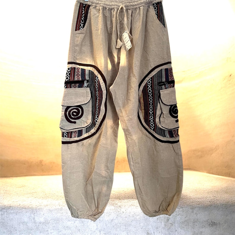 May include: Tan-colored, loose-fitting pants with a drawstring waist and two cargo pockets. The pockets and side panels feature a colorful, patterned design with red, blue, and black accents. The pants have a relaxed, bohemian style.