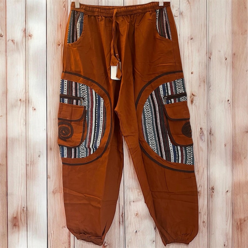 May include: Rust-colored cargo pants with a drawstring waist and pockets. The pants feature decorative panels with a mix of patterns in shades of brown, white, blue, and red. The pockets have a spiral design.