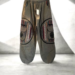 May include: Gray, loose-fitting pants with decorative patches. The pants feature two large, circular patches with colorful, patterned fabric and black trim. Each patch has a pocket and a spiral design. The pants have a drawstring waist.