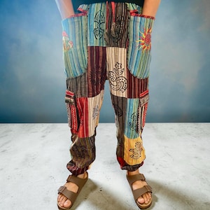 Peut inclure: Pantalon patchwork multicolore avec des motifs rayés, géométriques et solaires. Le pantalon est doté de grandes poches et est porté avec des sandales marron. Le style est bohème, décontracté.