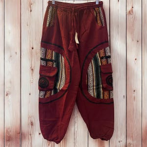 May include: Maroon cargo pants with decorative tribal-style fabric panels and pockets. The pants have a drawstring waist and are hanging against a wooden background. The panels feature black, white, yellow, and green patterns.