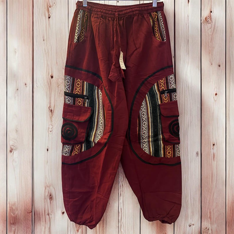 May include: Maroon cargo pants with decorative tribal-style fabric panels and pockets. The pants have a drawstring waist and are hanging against a wooden background. The panels feature black, white, yellow, and green patterns.