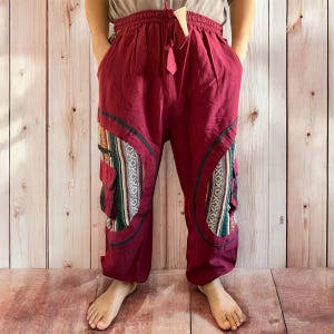 May include: Burgundy-colored pants with a drawstring waist and pockets. The pants feature decorative panels with a geometric pattern in shades of green, white, and black. The pants have a relaxed fit and are made of a lightweight fabric.
