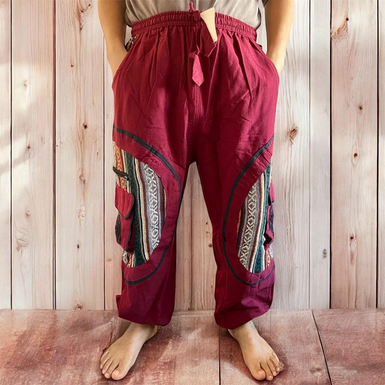 May include: Burgundy-colored pants with a drawstring waist and pockets. The pants feature decorative panels with a geometric pattern in shades of green, white, and black. The pants have a relaxed fit and are made of a lightweight fabric.
