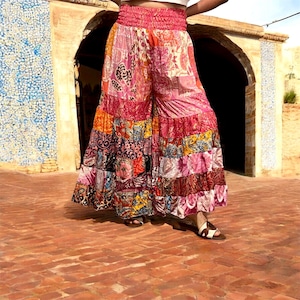 May include: Wide-leg pants featuring a vibrant patchwork design with various floral and paisley patterns in shades of pink, orange, yellow, and blue. The garment has a high-rise, elasticized smocked waistband and a tiered, flowing silhouette.