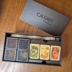 Op de afbeelding: Een zwarte Catan Travel box met een deksel voorzien van de in reliëf gedrukte tekst CATAN Travel box. De open doos bevat vier zwarte opbergbakjes met een rasterpatroon voor gekleurde fiches en drie stapels met geïllustreerde grondstoffenkaarten.