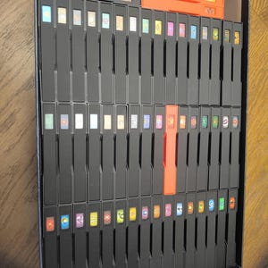 Puede incluir: Una caja de almacenamiento de juegos de mesa con numerosos porta tarjetas negros. Los porta tarjetas están dispuestos en filas y columnas, con cartas de juego coloridas visibles. También hay compartimentos de almacenamiento naranjas y negros. La caja está sobre una superficie de madera.