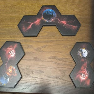 Puede incluir: Fichas hexagonales de tablero de juego con imágenes celestiales. Las fichas representan planetas, galaxias y rayos rojos sobre un fondo negro. Las fichas están dispuestas sobre una superficie de madera, lo que sugiere un juego de mesa.
