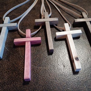 May include: A collection of six wooden cross pendants in various colors, including blue, pink, gray, and natural wood tones. Each cross is suspended from a light-colored cord, showcasing a simple, handcrafted design. The crosses are displayed on a dark, textured surface.