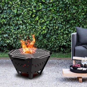 Puede incluir: Un brasero negro y geométrico con fuego encendido. El brasero tiene una parte superior cuadrada con una parrilla estampada y un cuerpo en forma de diamante. Las llamas son naranjas y amarillas. El brasero está sobre una superficie de grava, con un seto verde al fondo.