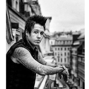 Può includere: Ritratto in bianco e nero di una persona con un'acconciatura mohawk, tatuaggi su entrambe le braccia e trucco scuro sugli occhi. La persona è appoggiata a una ringhiera di metallo, con uno sfondo urbano.