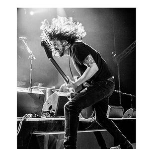 Puede incluir: Imagen en blanco y negro de un músico tocando la guitarra en el escenario. El músico lleva una camiseta negra y vaqueros, con pelo largo y un tatuaje en el brazo. La guitarra es oscura y el escenario está iluminado con luces brillantes.