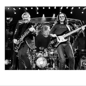 Pode incluir: Dois músicos tocando em um palco, um tocando uma guitarra elétrica preta e o outro tocando um baixo preto. O músico que toca a guitarra está usando um paletó preto e o músico que toca o baixo está usando uma camiseta preta com a palavra "RASI" impressa.