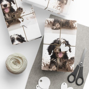 May include: Gift wrap featuring a playful black and white dog in a muddy puddle. The wrapping paper is tied with twine and adorned with a star-shaped tag. A spool of twine, scissors, and blank tags are also visible.