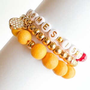May include: Three beaded bracelets. One bracelet has white beads spelling "HOME", another has yellow wooden beads and gold beads, and the third has white beads with the number "15". One bracelet has a gold-colored clasp with rhinestones.