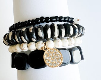 May include: A set of four bracelets featuring black and white beads. The bracelets are stacked on top of each other and include a charm with a gold-tone setting and clear rhinestones.