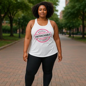 May include: A white tank top with the text "UNCOACHABLE" in a pink stamp design. The tank top is worn with black leggings. The image is taken outdoors with trees and a brick walkway in the background.