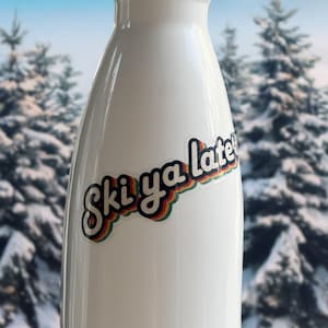 May include: White water bottle with a colorful "Ski ya later" sticker. The sticker has a retro design with rainbow stripes and a black outline. The background shows a snowy winter scene with evergreen trees.