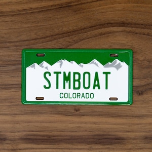 May include: A green and white license plate with the text "STMBOAT COLORADO". The license plate features a mountain range graphic at the top. The plate is rectangular with rounded corners and has three holes.