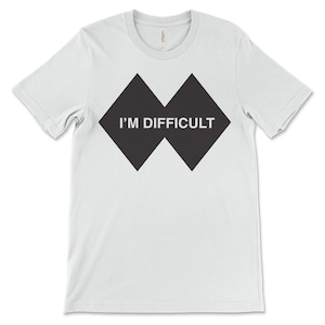 May include: White t-shirt with a black diamond graphic. The text "I'M DIFFICULT" is displayed in white within the graphic. The shirt has short sleeves and a crew neck. A casual, everyday garment.