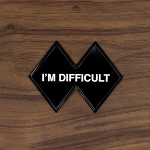 May include: A black, diamond-shaped emblem with the text "I'M DIFFICULT" in white lettering. The emblem is set against a brown, wood-grain background.