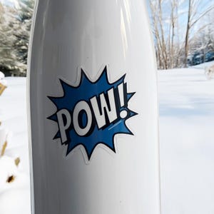 May include: White water bottle with a blue and white "POW!" comic book style sticker. The sticker is in a starburst shape with the word "POW!" in bold white letters. The background is a snowy outdoor scene.