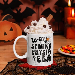 May include: White ceramic coffee mug with a black and white design that says "In my spooky physio era". The mug has a black spider, a ghost, a pumpkin, and a spider web on it. The mug is filled with whipped cream and chocolate chips and has a cinnamon stick sticking out of it. The mug is sitting on a black lace doily on a wooden table. There are black bats and orange candles in the background.