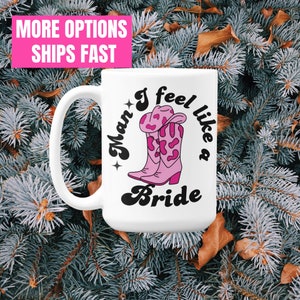 Könnte beinhalten: Eine weiße Keramiktasse mit einem schwarzen und rosafarbenen Design. Das Design zeigt ein Paar rosa Cowboystiefel mit einem weißen und rosafarbenen Muster und den Text "I feel like a Bride" in einer Retro-Schrift.