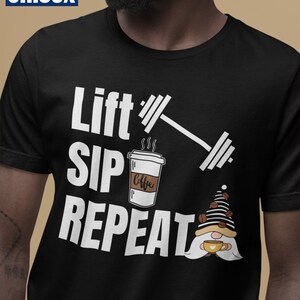 以下が含まれることがあります： 「Lift Sip Repeat」の白文字が入った黒色のユニセックスTシャツ。デザインにはバーベル、コーヒーカップ、コーヒーカップを持ったノームが含まれています。「Unisex」の文字が左上の青いボックスに入っています。