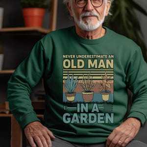 May include: A green sweatshirt with a vintage-style graphic that says "Never underestimate an old man in a garden". The graphic features three potted plants with a trowel.