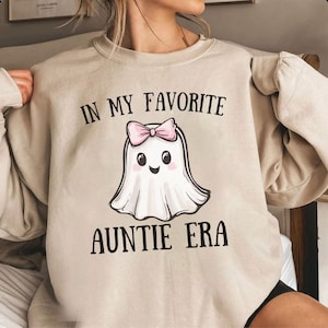 Pode incluir: Camisola bege com o texto "IN MY FAVORITE AUNTIE ERA" sobre um fantasma de desenho animado com um laço rosa. O fantasma tem um rosto sorridente e é delineado em preto. A camisola tem mangas compridas.