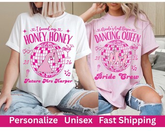 May include: Two t-shirts with pink graphics. One is white with "Honey Honey Ibiza" text and a bride illustration. The other is pink with "Dancing Queen Ibiza" text and dancing figures. Both have "20" and "26" dates.