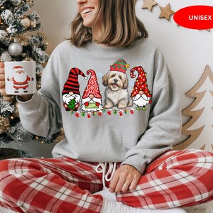 May include: A light grey unisex Christmas sweatshirt with a graphic of three gnomes and a dog wearing festive hats. The words "Merry Christmas" are displayed. The sweatshirt is paired with red and white plaid pajama bottoms.