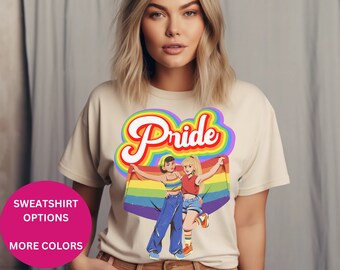Puede incluir: Una camiseta beige con un gráfico de arcoíris de dos mujeres sosteniendo una bandera arcoíris. El texto "Pride" está escrito en un arcoíris sobre las mujeres.