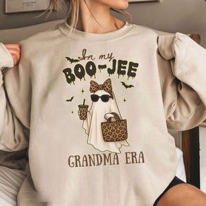 May include: Beige sweatshirt featuring a ghost graphic with sunglasses and a leopard print bow, holding a coffee cup and tote bag. The text reads "In my BOO-JEE Grandma ERA" with bats and stars.