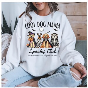 May include: A white sweatshirt featuring the words "COOL DOG MAMA" and a graphic of four dogs in Halloween costumes. The dogs are dressed as a witch, mummy, vampire, and skeleton. The words "Spooky Club" and "Very Demure, very Spooktacular" are below.
