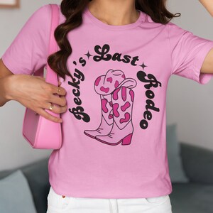 Pode incluir: Camiseta rosa com um gr&aacute;fico preto de uma bota de cowboy com um design de vaca rosa e branca. O texto "Becky's Last Rodeo" est&aacute; escrito em uma fonte cursiva preta ao redor da bota.