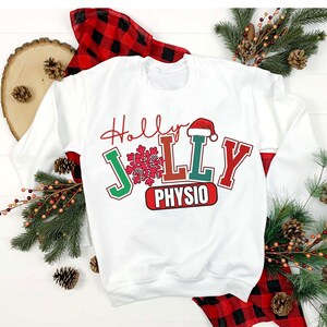 May include: White sweatshirt with the words "Holly Jolly Physio" in red, green, and white. The design includes a snowflake and a Santa hat. The sleeves have red plaid accents. Christmas-themed apparel.