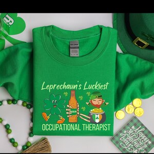 以下が含まれることがあります： 「Leprechaun's Luckiest Occupational Therapist」のテキストと聖パトリックの日のグラフィックが特徴の緑色のスウェットシャツ。デザインには、漫画のレプラコーン、四つ葉のクローバー、ビールのボトル、金のポットが含まれています。