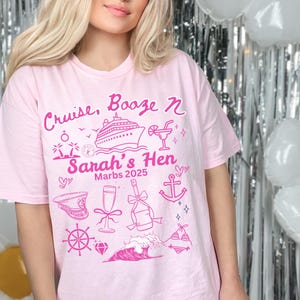 Op de afbeelding: Roze T-shirt met de tekst "Cruise, Booze & Sarah's Hen" en een cruiseschip, palmbomen, een cocktail, een anker, een scheepswiel, een diamant, een golf en een badpak. De tekst "Marbs 2025" staat ook op het shirt.