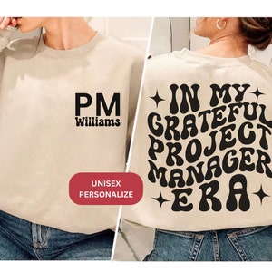 May include: A beige sweatshirt with the text "PM Williams" on the front and "In My Grateful Project Manager Era" on the back. The text is in a retro, wavy font.