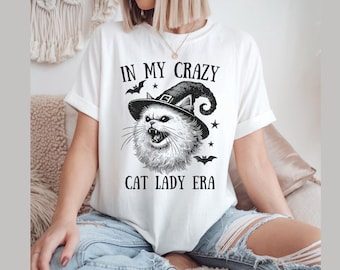 Pode incluir: Camiseta branca com uma ilustração em preto e branco de um gato usando um chapéu de bruxa. O texto "IN MY CRAZY CAT LADY ERA" é impresso acima e abaixo do gráfico do gato. A camisa tem mangas curtas.