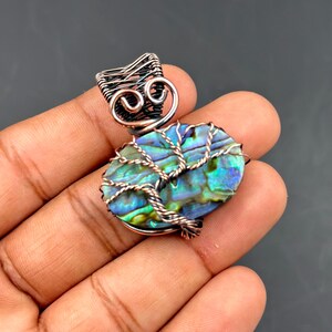 Op de afbeelding: Abalone schelpen hanger met een koperdraad boom ontwerp. De iriserende blauwe en groene schelp is omhuld met gedraaide koperdraad, met een geweven koperen bale. De hanger wordt in een hand tegen een donkere achtergrond gehouden.